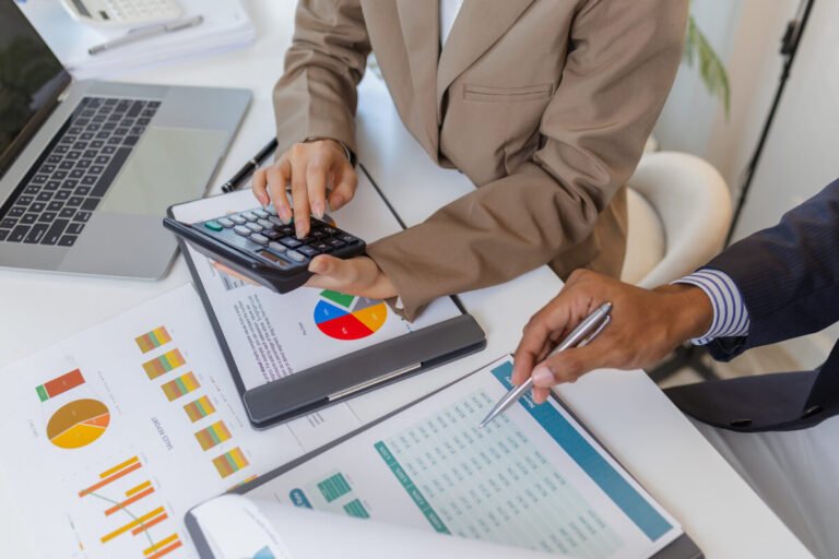 Accountant using a calculator while reviewing sales reports and financial charts with a colleague during a business consultancy meeting.