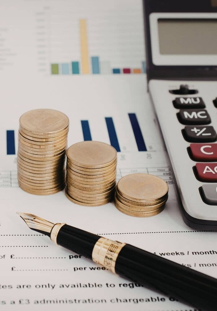 Stacks of British pound coins next to a calculator and fountain pen on a payroll document with financial charts in the background.
